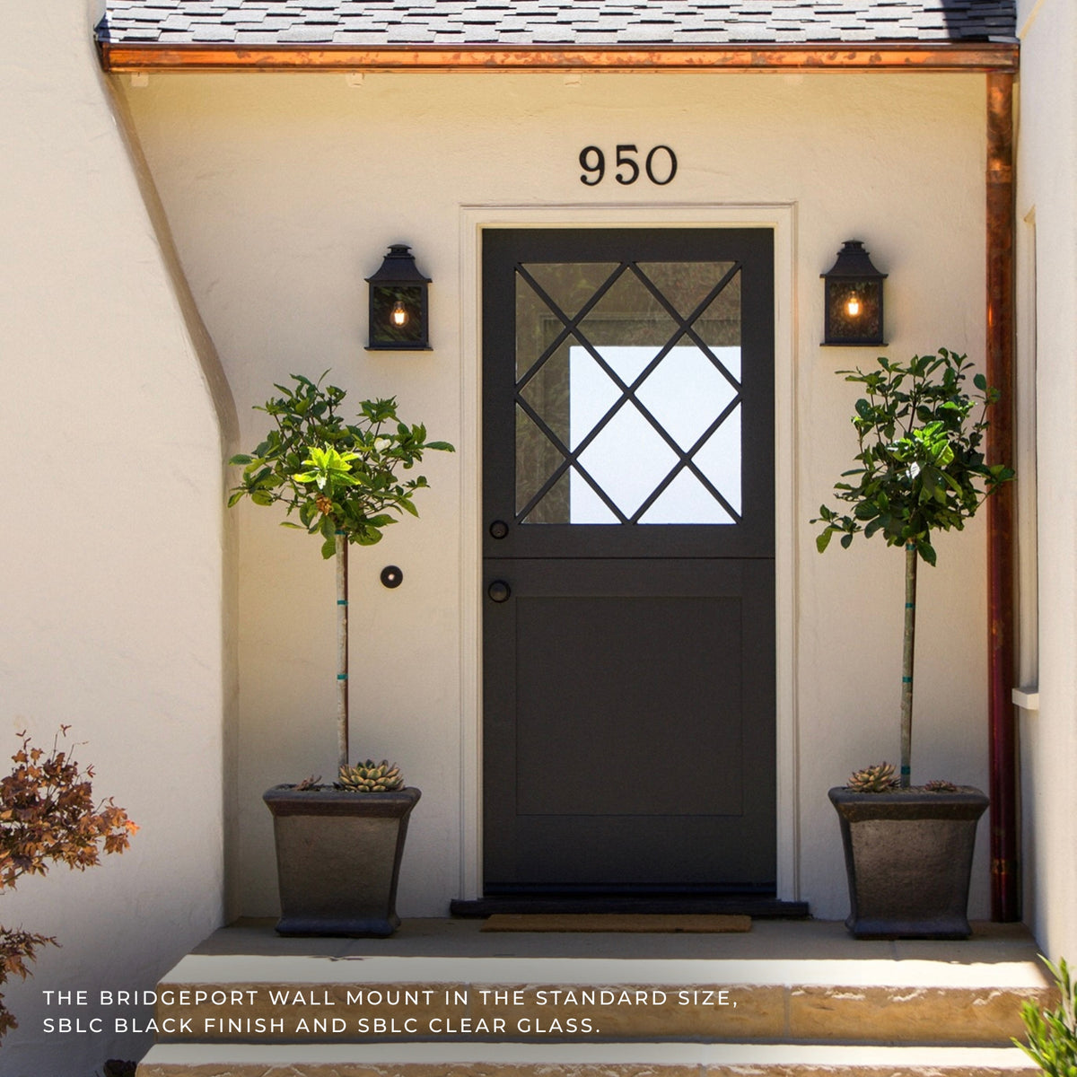 A black exterior wall-mounted door light with clear glass and a diamond crossbar pattern, shown on a beige stucco door frame next to potted plants. The text at the bottom reads 'THE BRIDGEPORT WALL MOUNT IN THE STANDARD SIZE, SBLC BLACK FINISH AND SBLC CLEAR GLASS.