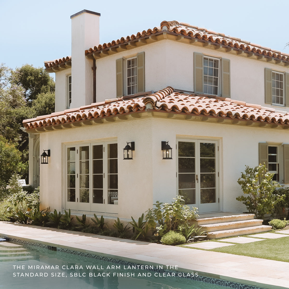 A stucco house exterior with terracotta tile roofs, shutters, outdoor wall lanterns, a pool edge, and landscaping. Text at the bottom reads: 'THE MIRAMAR CLARA WALL ARM LANTERN IN THE STANDARD SIZE, SBLC BLACK FINISH AND CLEAR GLASS.
