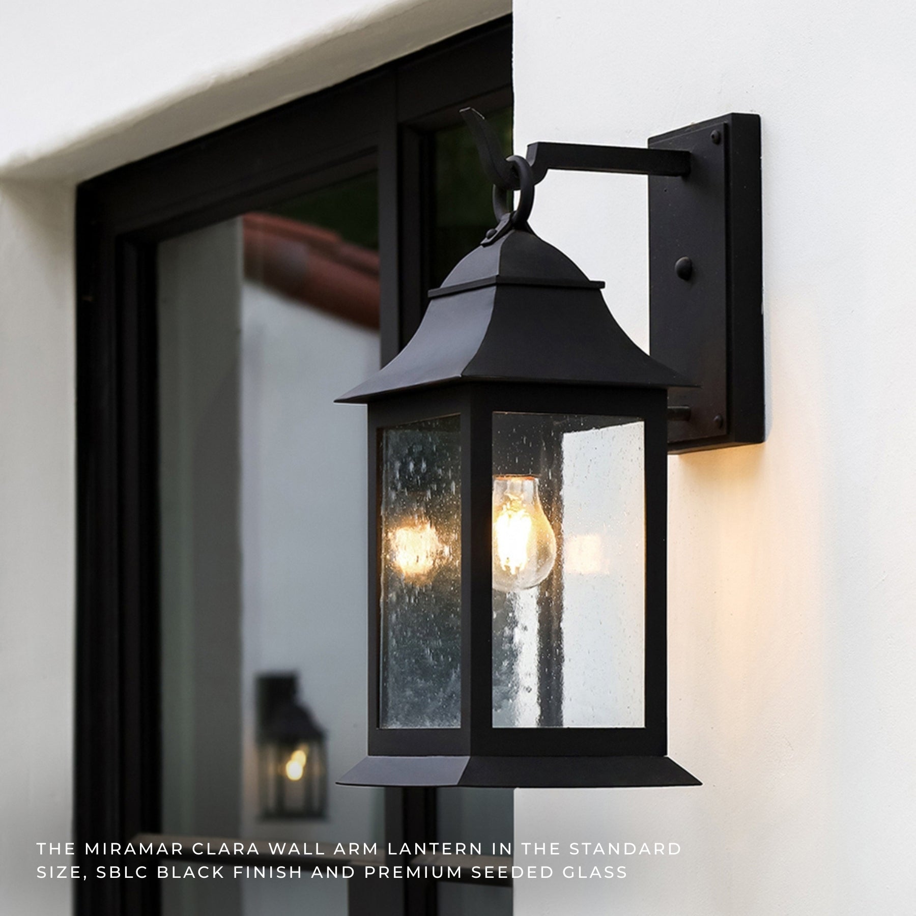 Black Miriam Clara outdoor wall lantern mounted on a white exterior wall next to a black-framed window, with the warm glow of the exposed light bulb visible through the seeded glass.