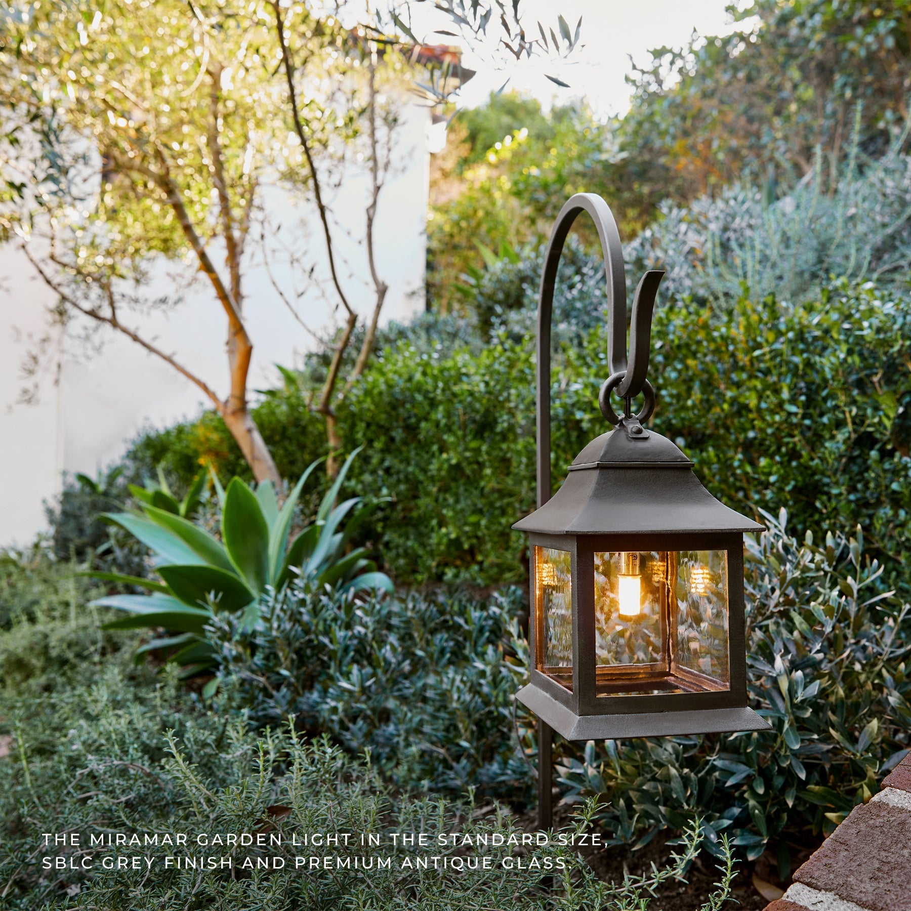 A classic outdoor garden lantern light with a curved hanging arm, shown lit in a lush garden setting with green foliage and plants. The lantern has a dark grey antique metal finish with clear glass panels.