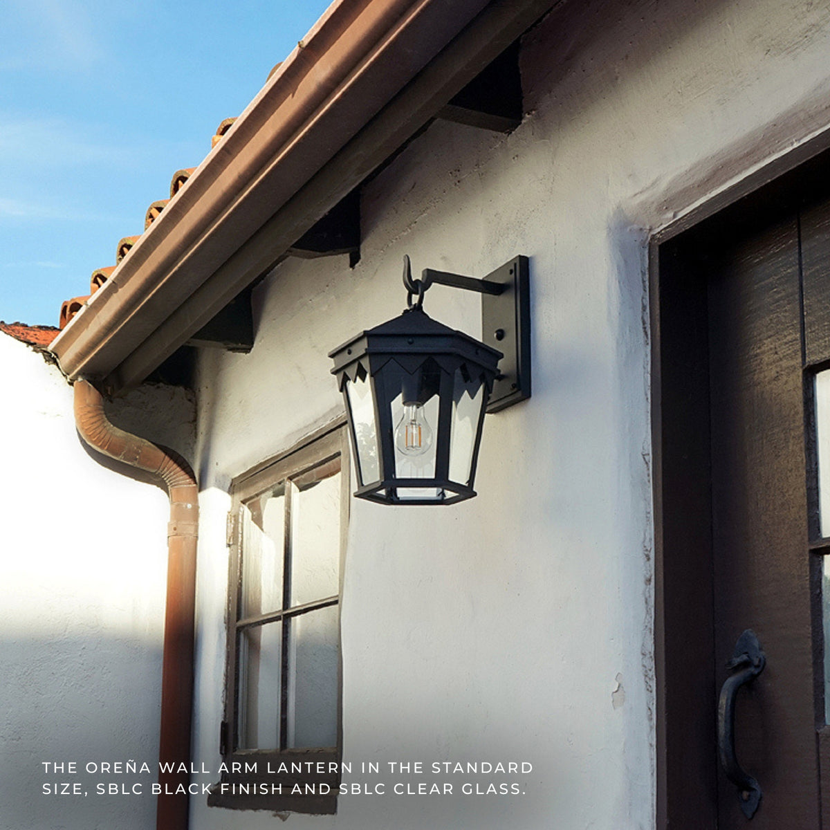 Black outdoor wall lantern mounted on a white stucco house exterior next to a dark wooden door, with roofline and gutter visible above.