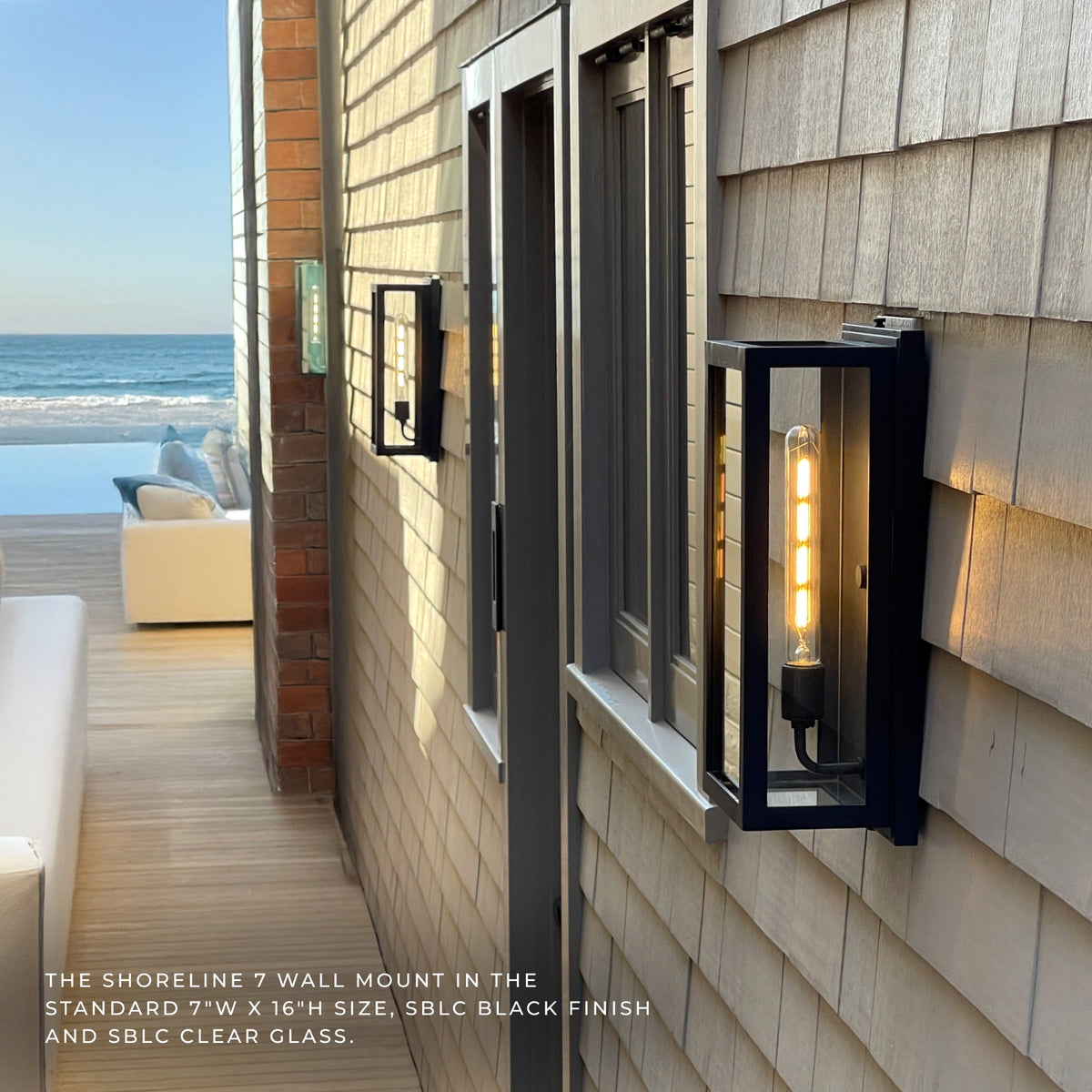 A black outdoor wall sconce with a clear glass front and visible warm Edison-style bulb, mounted on a beige shingled house exterior beside a deck and ocean view. Text at the bottom identifies the Shoreline 7 wall mount and describes the finish and glass.