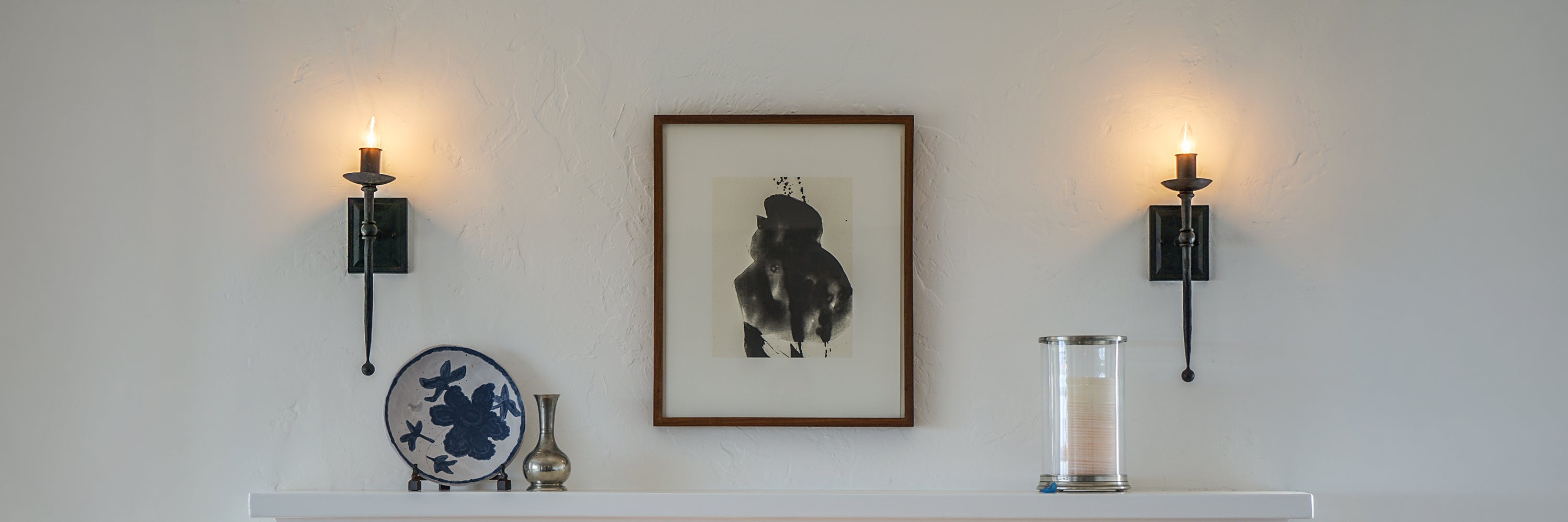 A framed black and white art print hangs on a white wall between two black wall sconces, above a white shelf holding a blue-and-white decorative plate, a small vase, and a glass candle holder.