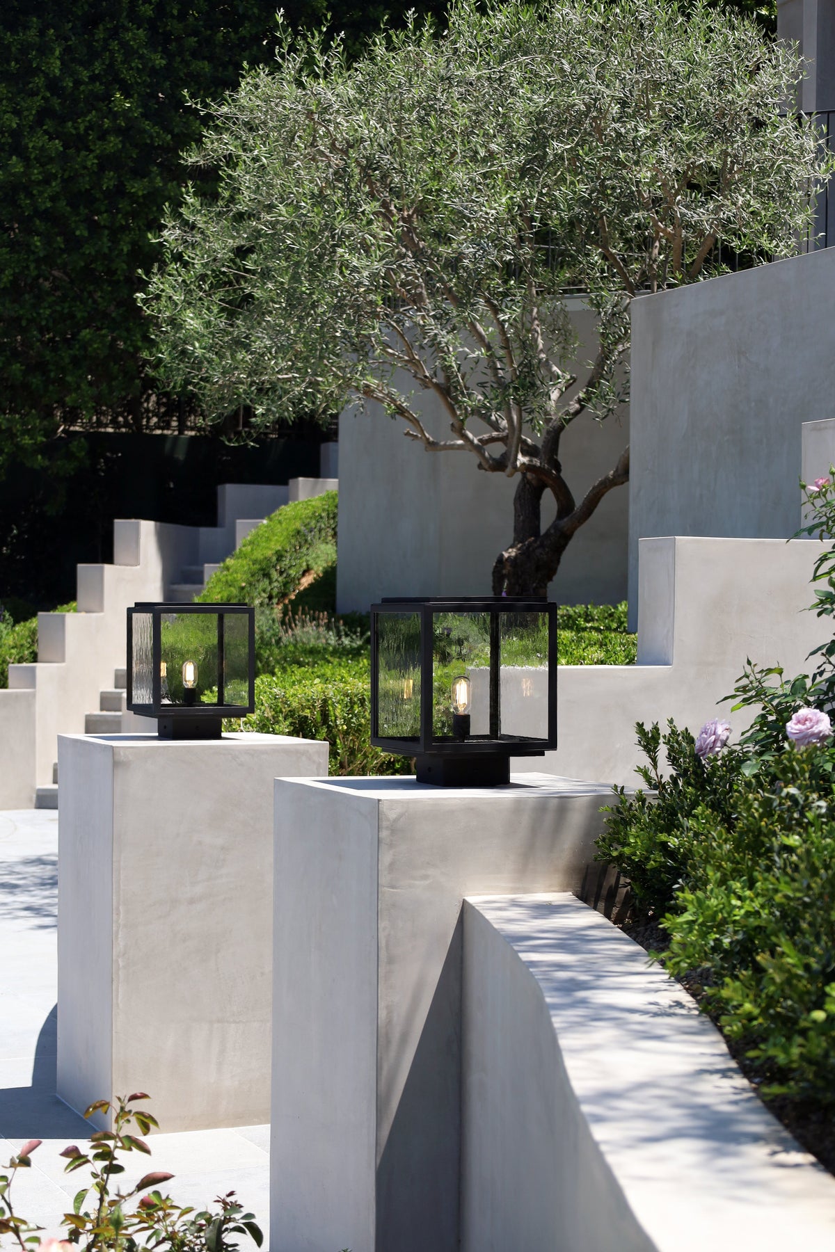 Decorative outdoor setting with black lanterns on white stone plinths, surrounded by greenery.