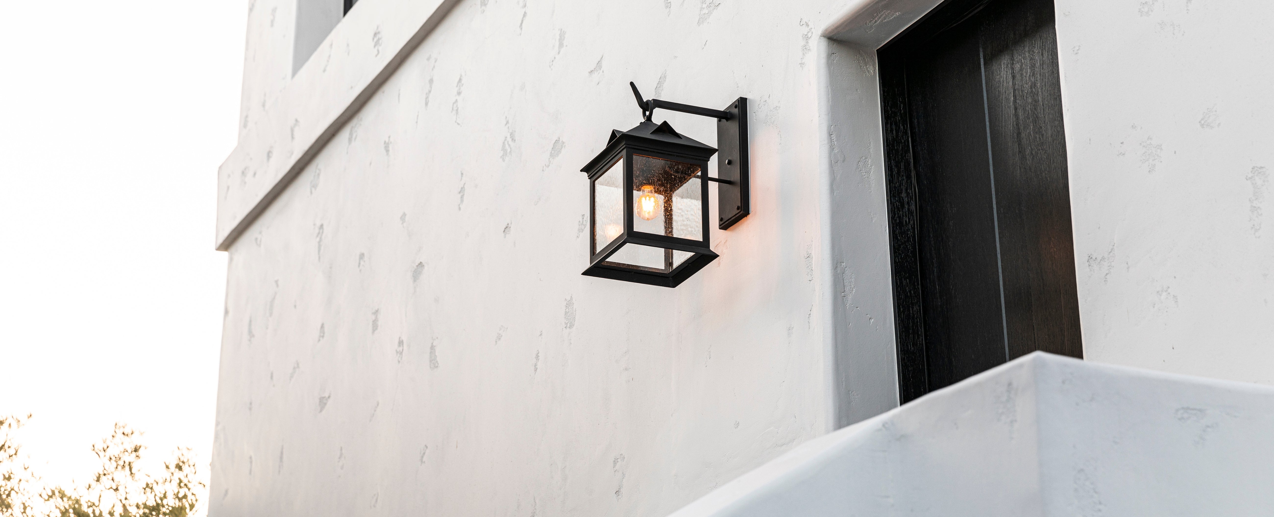 A black outdoor wall sconce with a warm glowing light, mounted on a white stucco exterior wall next to a dark door.