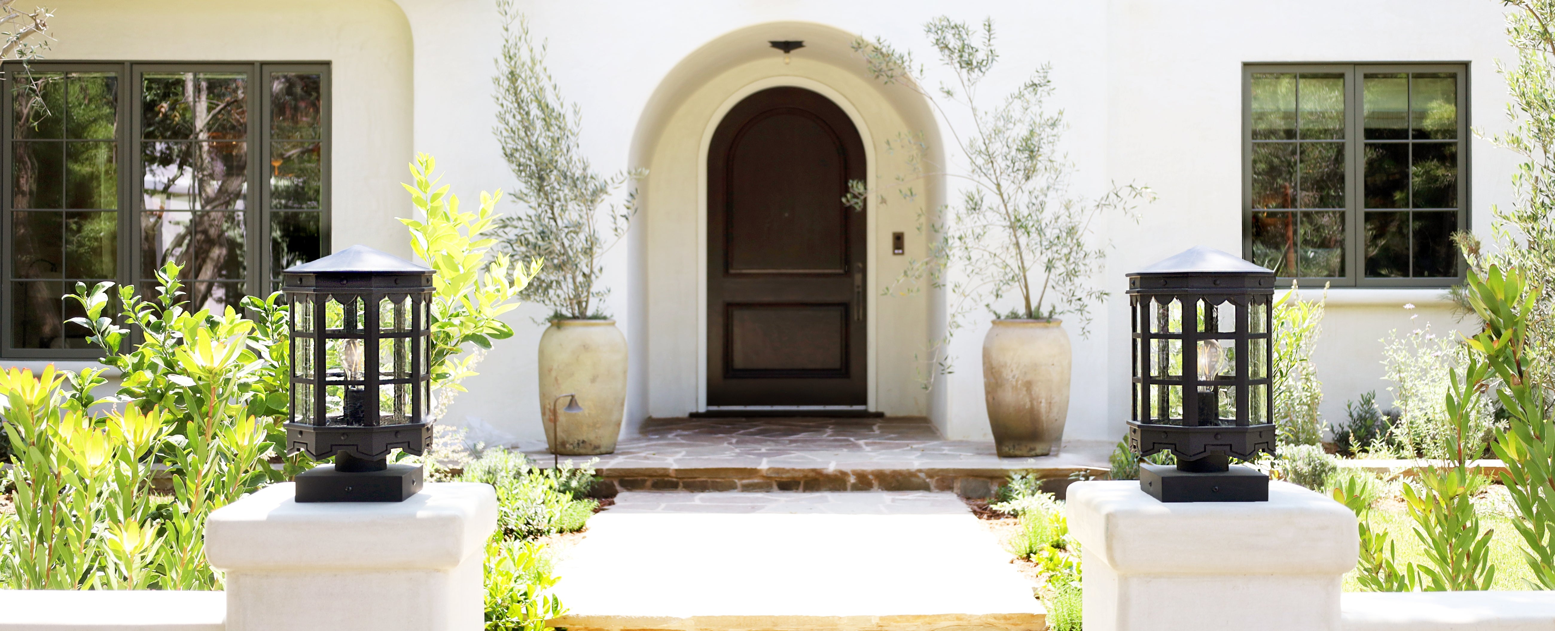 A symmetrical outdoor view of a white stucco house entrance with an arched dark brown door, flanked by large beige planters with olive trees. Two black lantern-style outdoor lights sit on white pedestals in the foreground, surrounded by green plants.