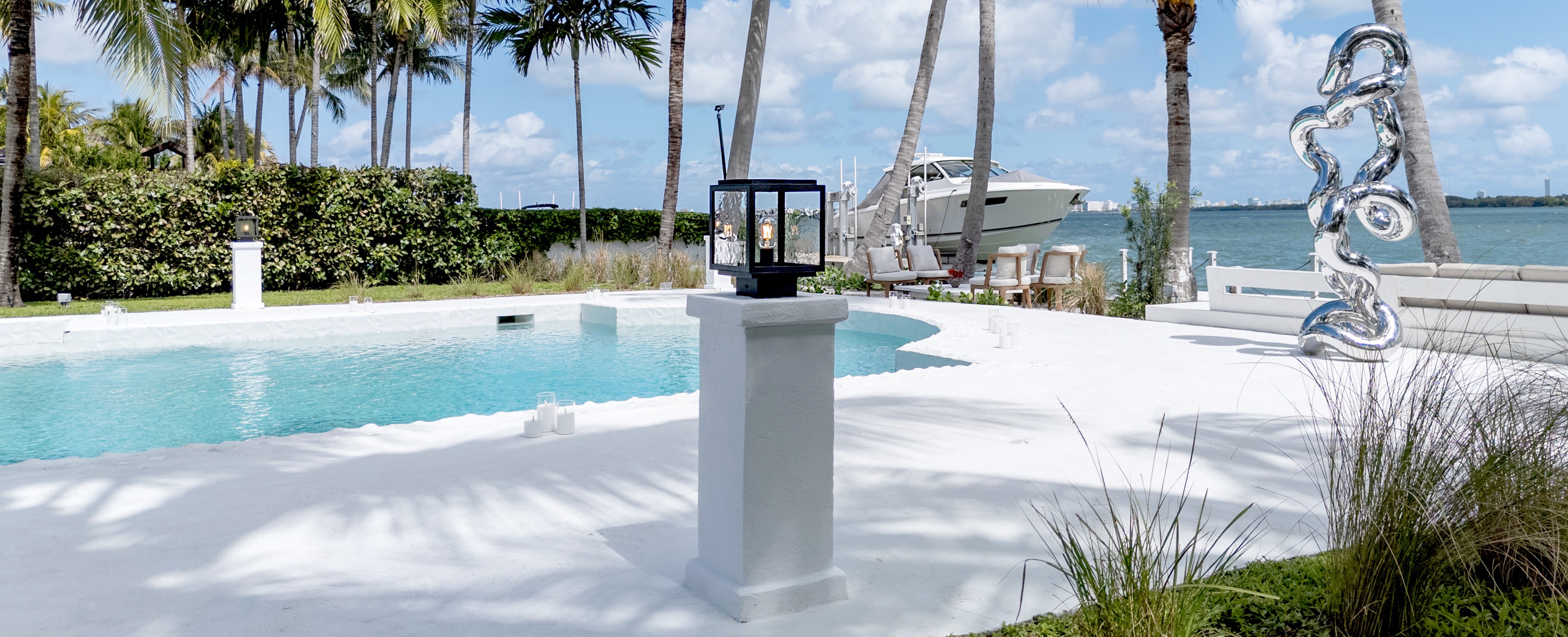 A white outdoor pedestal lamp with a black glass shade stands on a white patio beside a swimming pool, with palm trees, water, and a boat in the background.