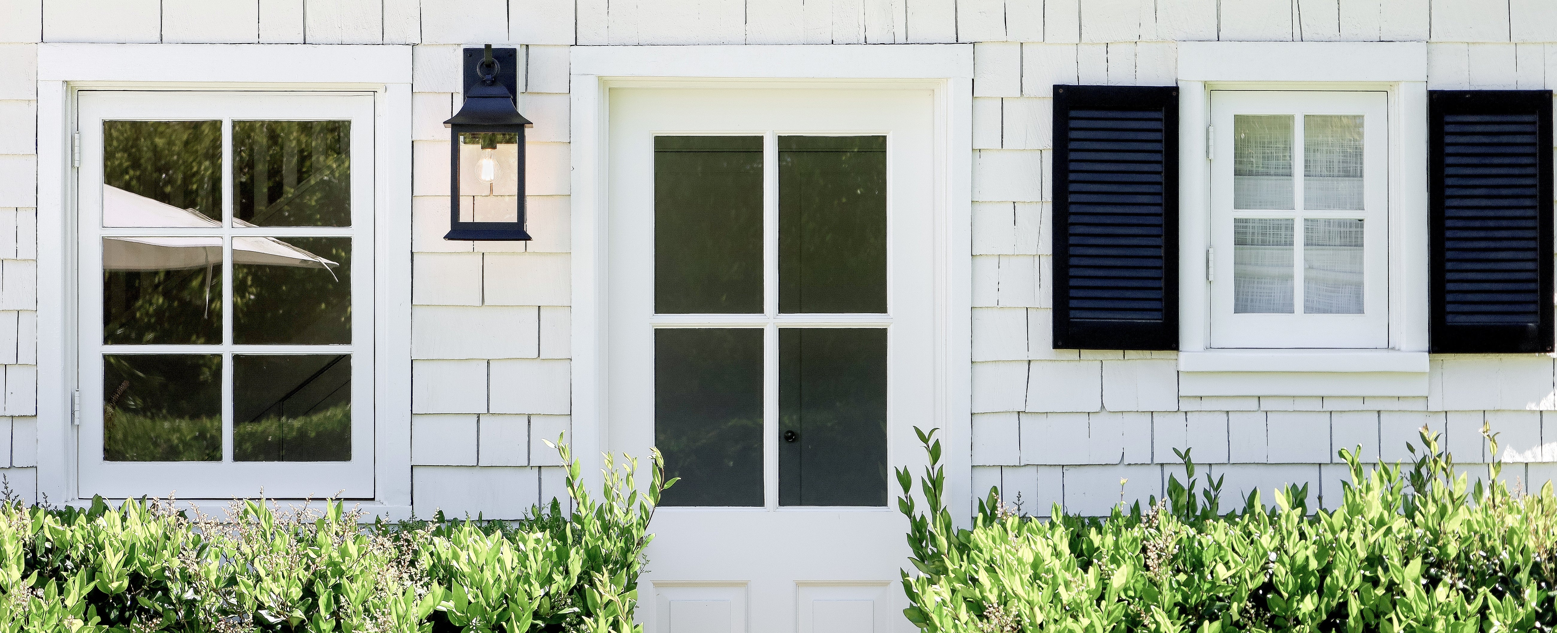 A white shingled house exterior with a white front door, black shutters, a black wall lantern, and green shrubs in the foreground.
