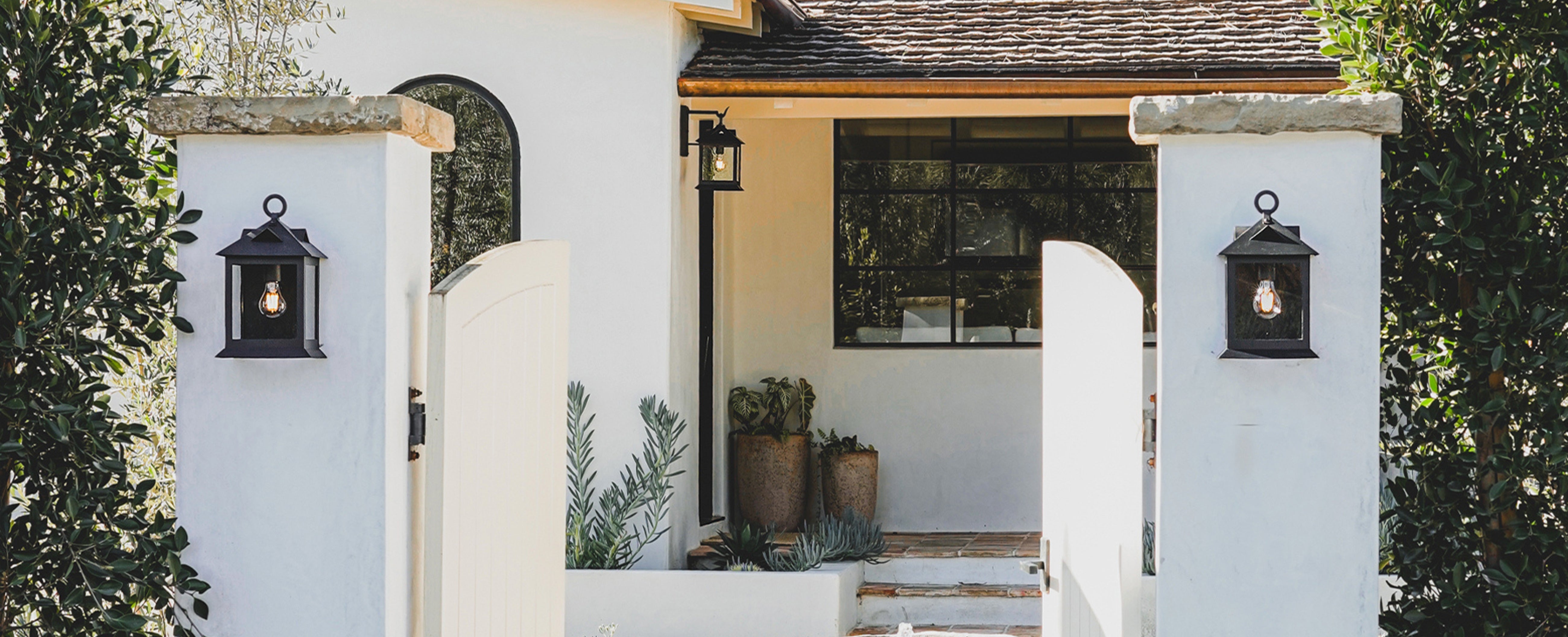 A white stucco house entrance with two black lantern-style wall sconces mounted on stone pillars flanking an open white gate. The scene includes a covered entryway, dark window framing, potted plants, and surrounding greenery.