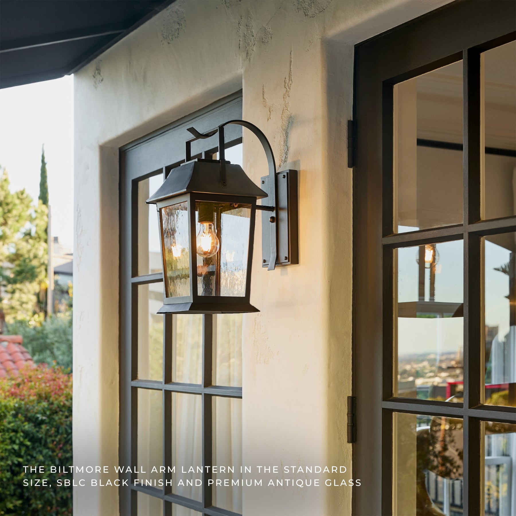 Biltmore wall arm lantern outdoor light fixture mounted on a stucco wall next to a dark-framed glass door, with warm light glowing from the bulb. Text at the bottom reads: THE BILTMORE WALL ARM LANTERN IN THE STANDARD SIZE, SBLC BLACK FINISH AND PREMIUM ANTIQUE GLASS.