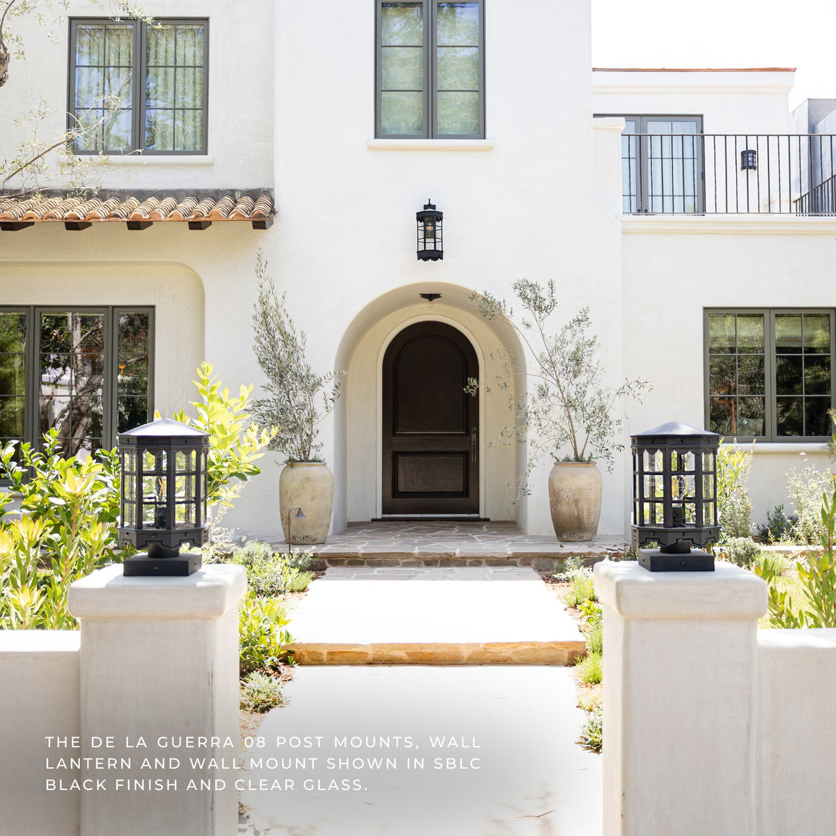 A white stucco house facade with an arched dark front door, black wall lanterns mounted above the door and on stone posts in the foreground, potted plants, windows, and a walkway leading to the entrance. Text at the bottom reads: 'THE DE LA GUERRA OR POST MOUNTS, WALL LANTERN AND WALL MOUNT SHOWN IN SBC BLACK FINISH AND CLEAR GLASS.
