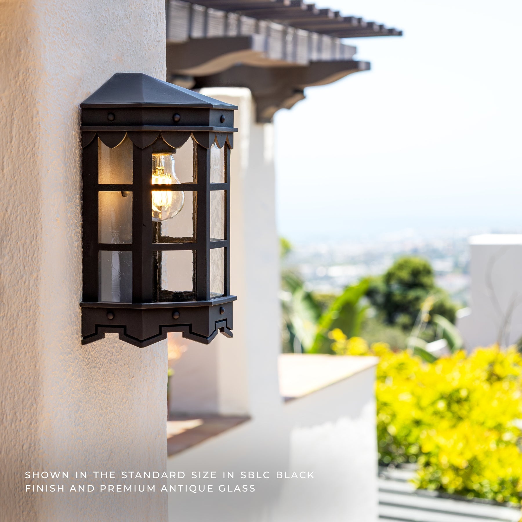 A black outdoor wall lantern light fixture mounted on a white stucco house pillar, with a glowing bulb inside and a blurred outdoor cityscape background. Text at the bottom reads: 'SHOWN IN THE STANDARD SIZE IN SBC BLACK FINISH AND PREMIUM ANTIQUE GLASS'.