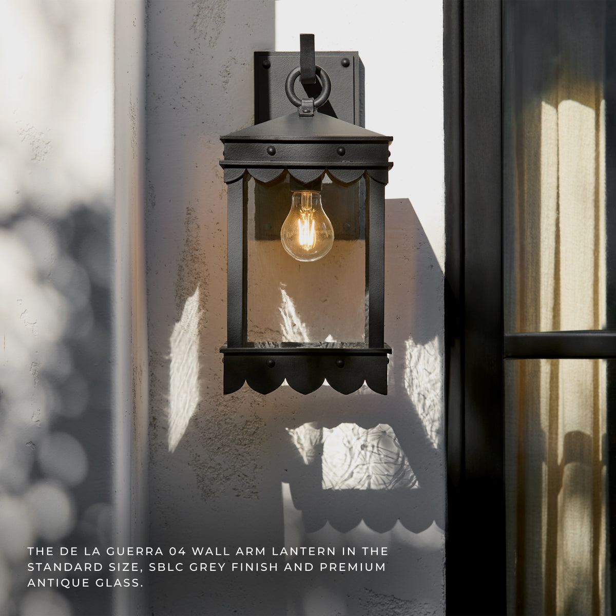 Black outdoor wall lantern mounted on a textured stucco wall next to a glass door, with a warm glowing bulb inside. Text at the bottom reads: 'THE DE LA GUERRA 04 WALL ARM LANTERN IN THE STANDARD SIZE, SBLC GREY FINISH AND PREMIUM ANTIQUE GLASS.