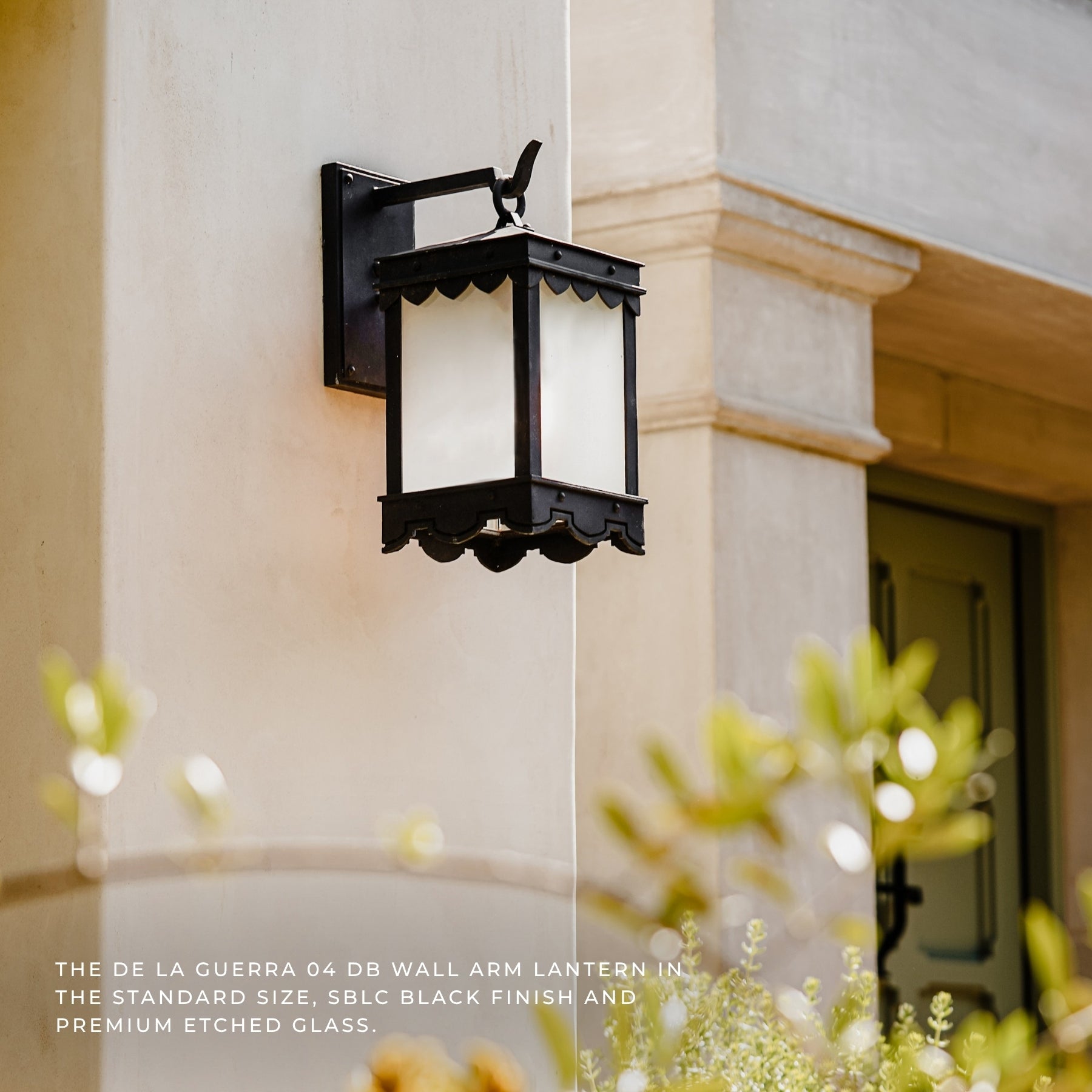 A black outdoor wall lantern with frosted white glass panels mounted on a beige stucco building wall, with blurred greenery in the foreground and text at the bottom describing the lantern.