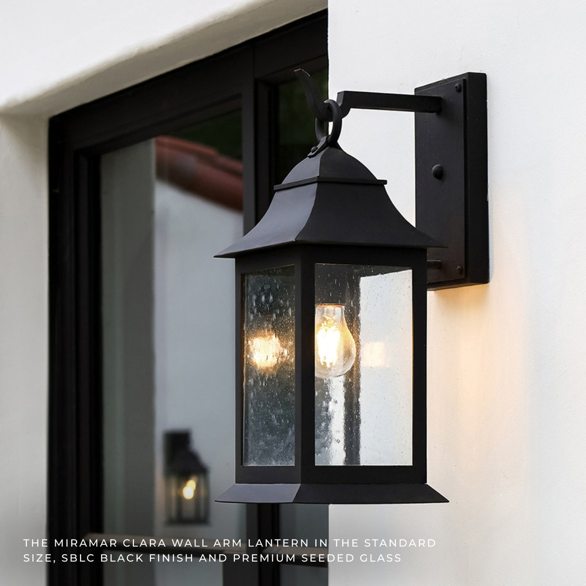 Black Miriam Clara outdoor wall lantern mounted on a white exterior wall next to a black-framed window, with the warm glow of the exposed light bulb visible through the seeded glass.