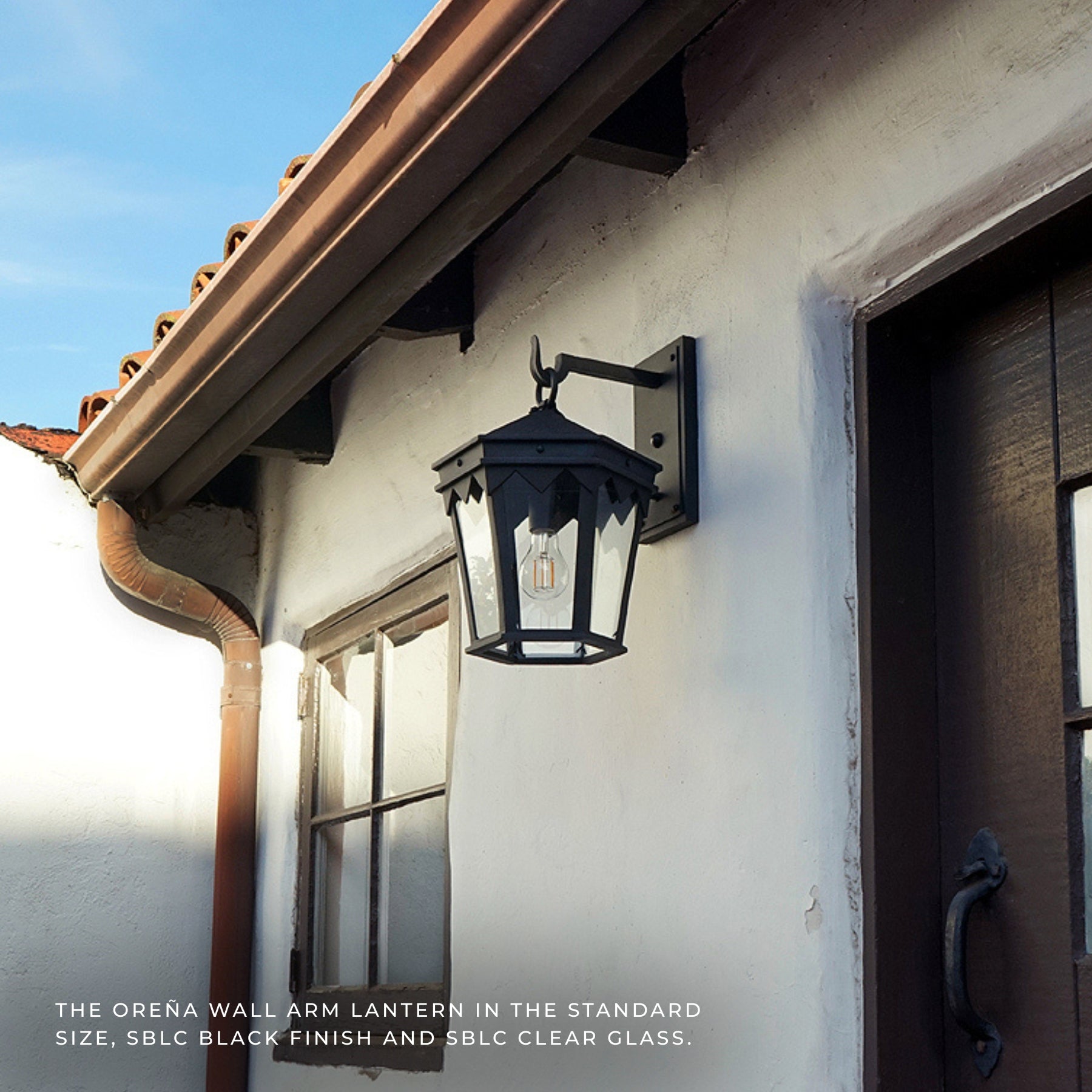 Black outdoor wall lantern mounted on a white stucco house exterior next to a dark wooden door, with roofline and gutter visible above.