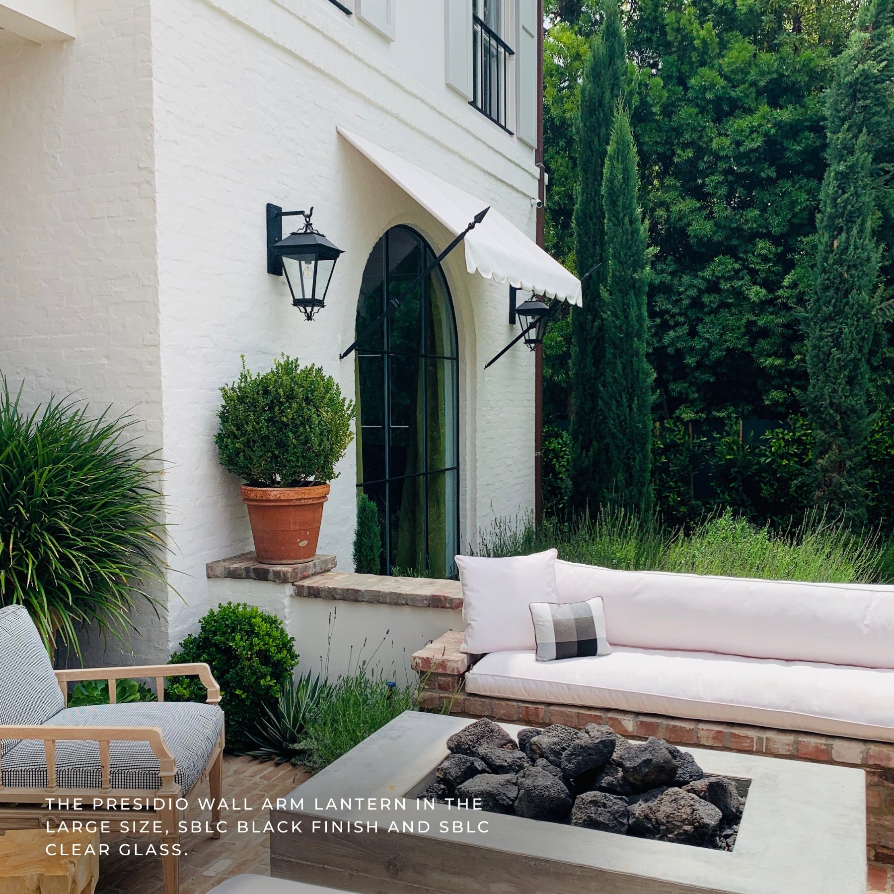 Outdoor patio scene beside a white stucco house with a black Presidio wall arm lantern mounted on the wall. The setting includes a light pink outdoor sofa, a fire pit with dark rocks, green plants, tall cypress trees, and a garden chair.