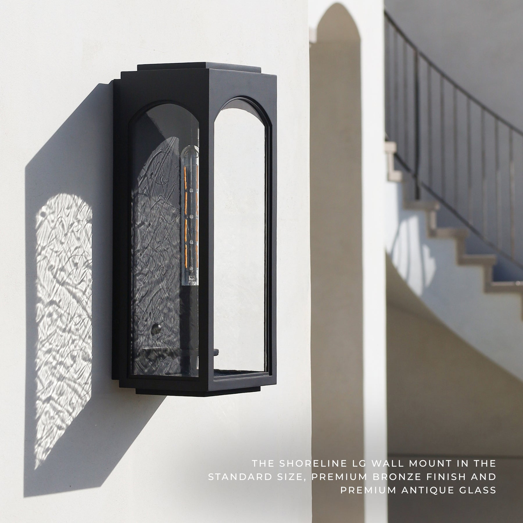 A black Shoreline LG wall mount light fixture with textured glass, mounted on a white exterior wall casting a shadow, with a staircase in the background.