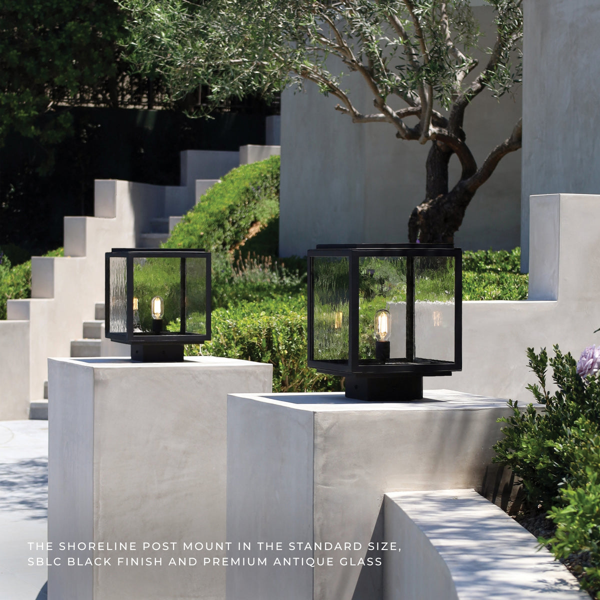 Two black outdoor lantern-style post lights mounted on white stone pedestals in a landscaped garden with steps, greenery, and a tree. Text at the bottom describes the shoreline post mount and materials.