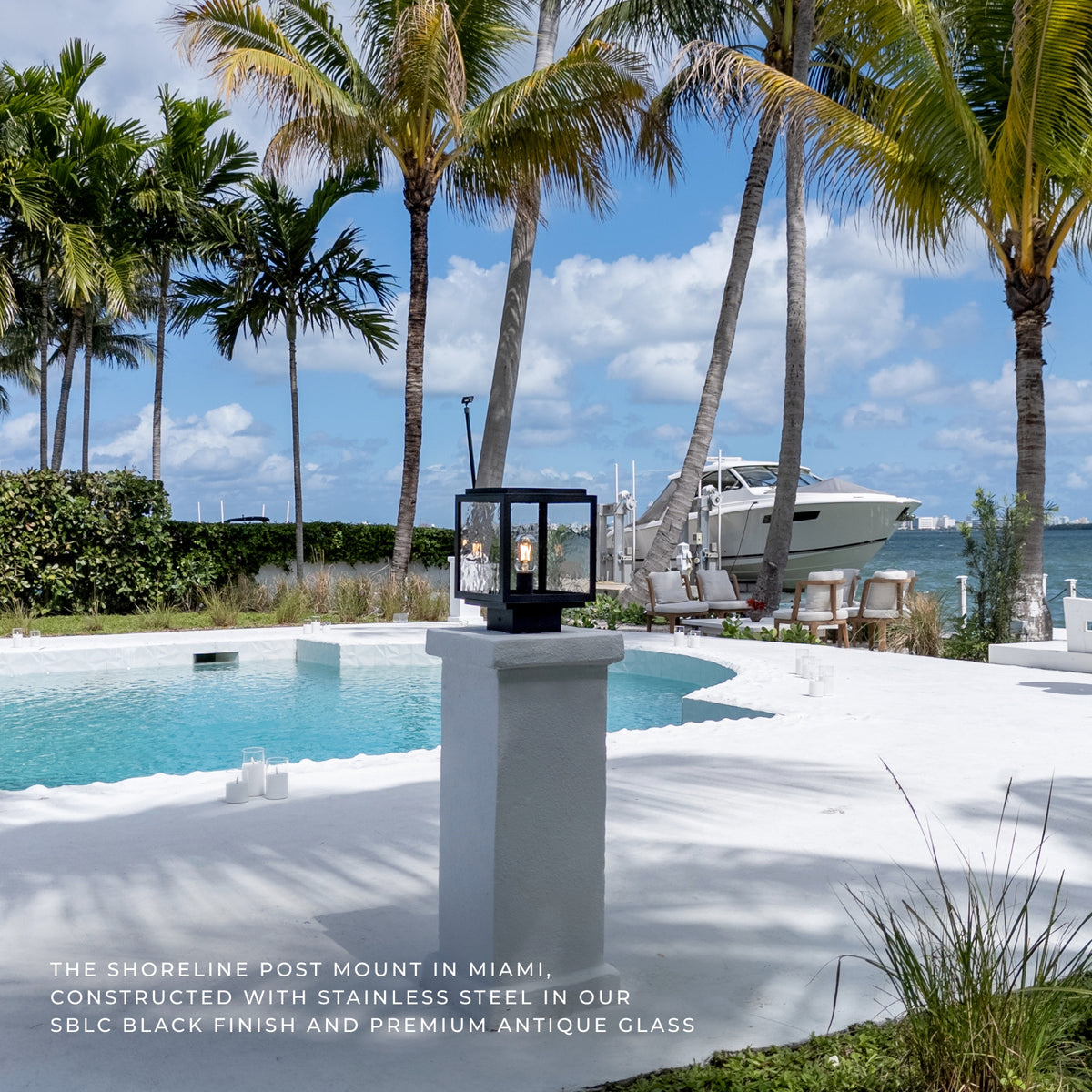 Outdoor post-mounted lantern light with black stainless steel framing and antique glass, shown on a white poolside pedestal in a tropical Miami setting with palm trees, water, and a yacht.