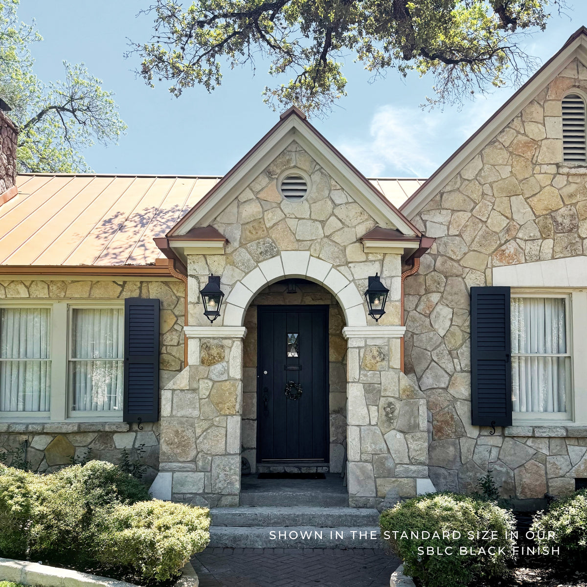 A front-facing view of a stone house entrance with a black door, black shutters, and a copper roof under a blue sky. Text at the bottom reads 'Shown in the standard size in our SBLC black finish.